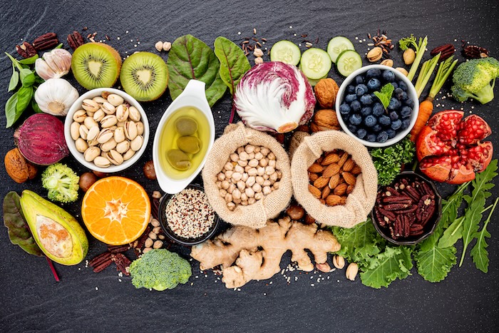 Vielfältige Auswahl an pflanzlichen Lebensmitteln auf schwarzem Untergrund, darunter Kiwi, Knoblauch, Pistazien, Olivenöl, Radicchio, Blaubeeren, Granatapfel, Brokkoli, Avocado, Orange, Quinoa, Kichererbsen, Mandeln, Pekannüsse, Ingwer, Gurkenscheiben, Karotten, Rote Bete und frische Kräuter.